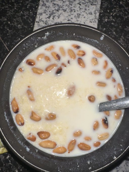 A bowl of creamy soup or porridge with peanuts floating on top and a spoon resting inside, placed on a dark surface with a granite-like pattern.