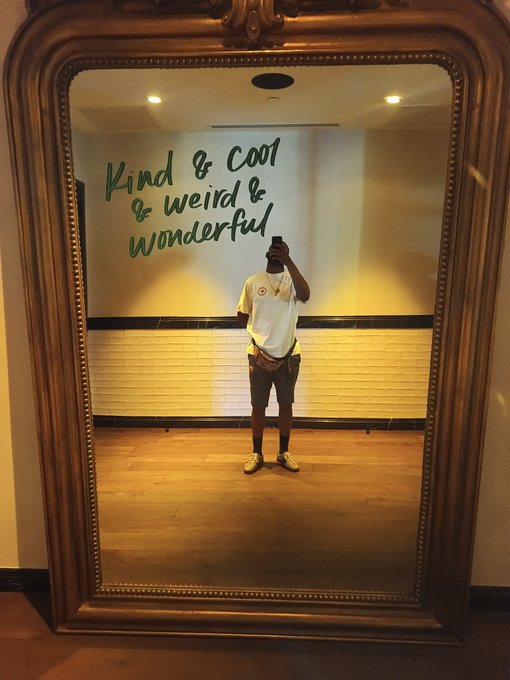 A person takes a mirror selfie in a large ornate gold frame, with the text 'Kind & cool & weird & wonderful' written in green on the wall behind them.
