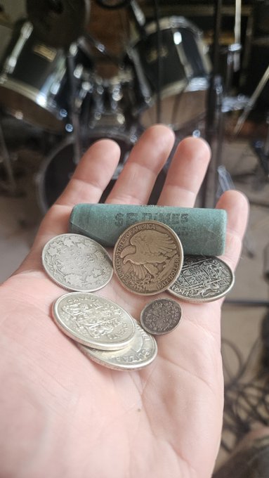 A hand cradles several silver coins and a rolled bill, with the word "MONIES" visible on the roll, alongside a drum set—embodying the sentiment "If you don't hold it, you don't own it." The tactile possession of physical currency reinforces the message that true ownership requires holding assets in hand, not just on paper.