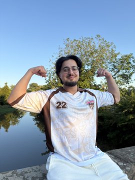 A man wearing glasses and a white sports jersey with the number "22" flexing his biceps outdoors near water and trees.