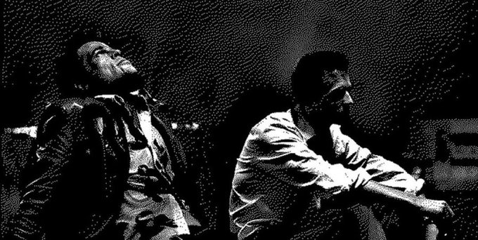 A high-contrast black and white image showing two men sitting together, one leaning back with his eyes closed while the other leans forward, both appearing contemplative.