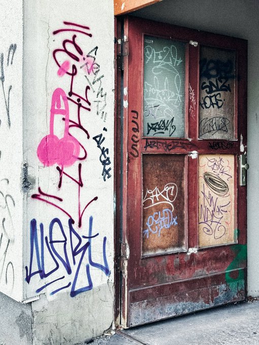 A weathered red door and adjacent concrete wall covered in colorful graffiti tags, including purple and black scribbles and a pink spray-painted figure.