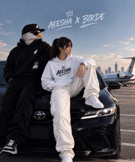 A promotional photo shows a person wearing an eagle mask and black clothing standing next to a woman in a white 'AEESHA' hoodie and cargo pants, leaning against a black Toyota car with an airplane in the background.