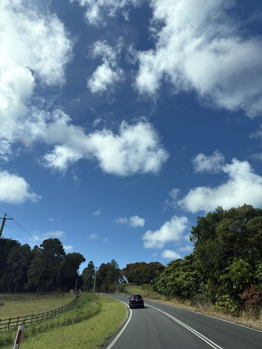 A scenic photograph of a car driving down a winding country road, flanked by green fields, trees, and a bright blue sky filled with scattered white clouds.