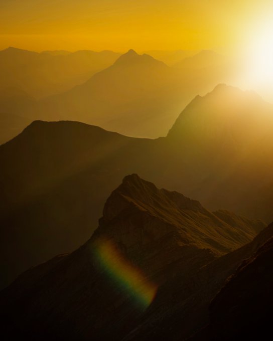 A scenic view of mountain peaks at golden hour with the sun glowing brightly and a rainbow lens flare visible in the foreground.
