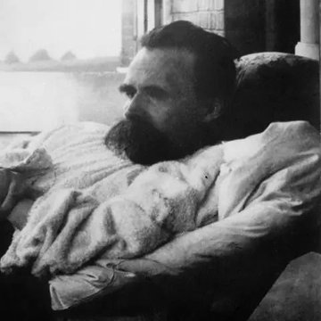 A vintage black-and-white photograph of a bearded man resting in bed, covered with a blanket, looking off to the side.