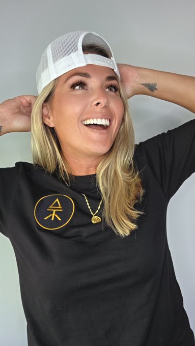 A smiling blonde woman wearing a white trucker cap backwards and a black t-shirt featuring a gold circular logo with a stylized tree/figure design, holding the cap with both hands, relates to the post "Check out @ATM_xrpl" as her apparel promotes the ATM brand associated with the XRP Ledger cryptocurrency.