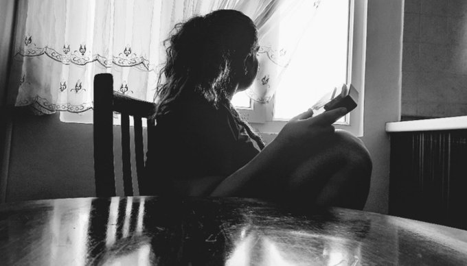 A black and white silhouette shows a young person sitting in a chair by a window with lace curtains, reading a small book.