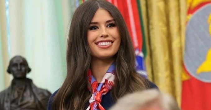 A smiling young woman with long brown hair, wearing a blue top and a patterned silk scarf, standing in front of a George Washington bust and a flag.