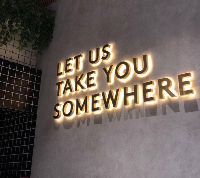 This image features an electric sign with the message "Let us take you somewhere" written in large letters. The sign has a lighted appearance that makes it stand out against the dark background. It's positioned above a white wall, creating a striking contrast between the sign and the wall. The words on the sign are clearly visible to everyone passing by. The sign seems to be an advertisement or a promotional message for something related to finance or investments, as suggested by the caption below it.