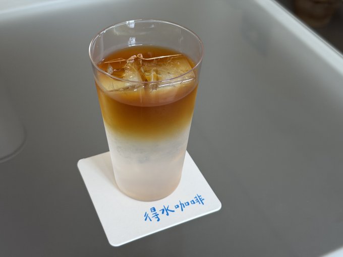 A layered iced coffee drink in a clear plastic cup with ice, sitting on a white coaster with the Chinese text '得水咖啡' (De Shui Coffee) in blue.