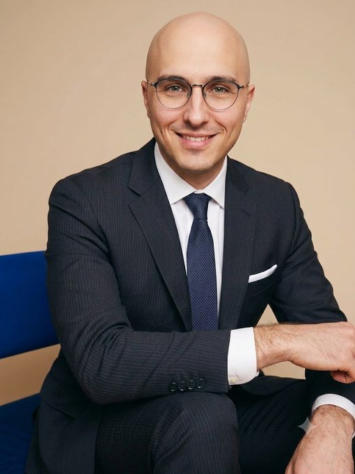 A professional portrait of a bald man with glasses, wearing a dark pinstripe suit, white shirt, and navy blue tie, smiling against a beige background.