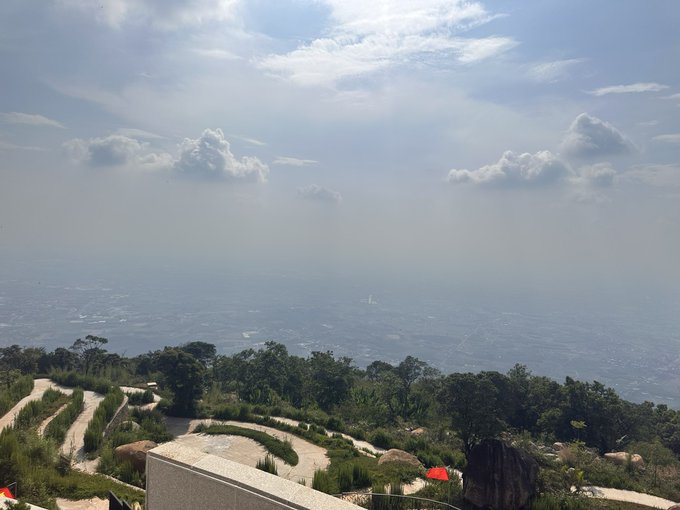 Scenic mountain overlook photograph showing a vast, hazy cityscape below with scattered clouds and sun rays piercing through the sky.