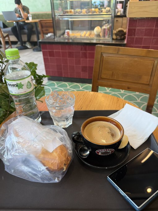 A tray of food and drinks at a cafe, including a bottle of Essent water, a glass of water, a coffee in a Johnny's cafe cup, a pastry in a plastic bag, and a smartphone, with a wooden chair nearby.