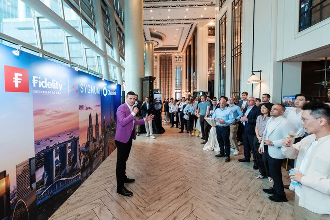 A man in a purple jacket speaks into a microphone at a corporate event, with banners for 'Fidelity International', 'SYGNUM', and 'Oktoink' visible behind him as attendees watch.