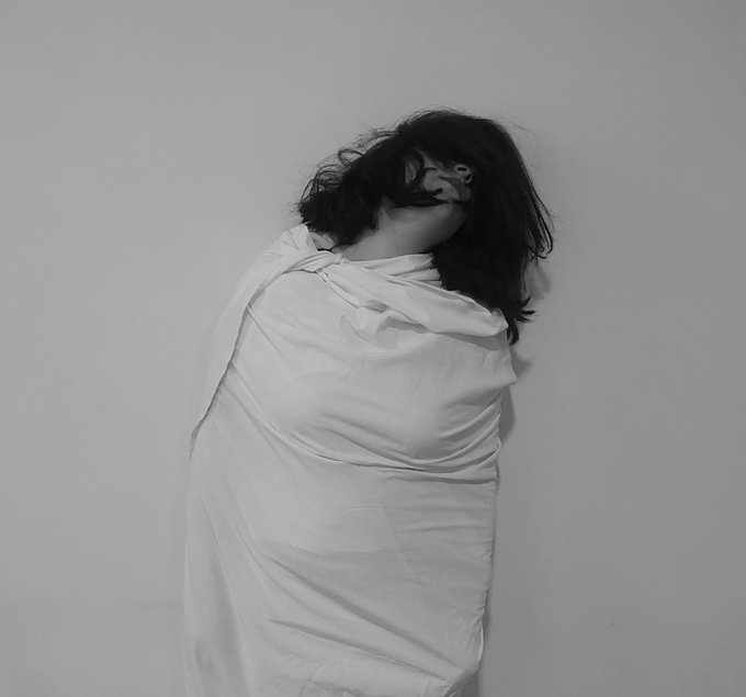 A black and white photograph of a person wrapped in a white sheet against a plain wall, with dark hair obscuring their face.