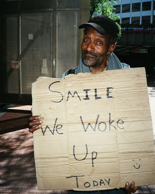 An older man with a beard holds a handmade cardboard sign that reads 'SMILE We Woke Up' with a smiley face, standing on a city sidewalk.