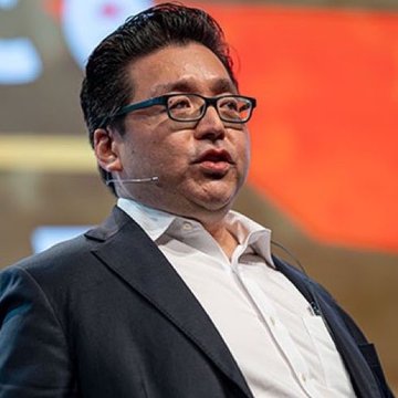 The image features a man wearing glasses and a suit jacket, giving him an intellectual appearance as he speaks at an event or conference. The background of the photo displays some electronic equipment that seems related to the Bitcoin market, adding to the overall atmosphere of the discussion about blockchain technology and cryptocurrency markets.