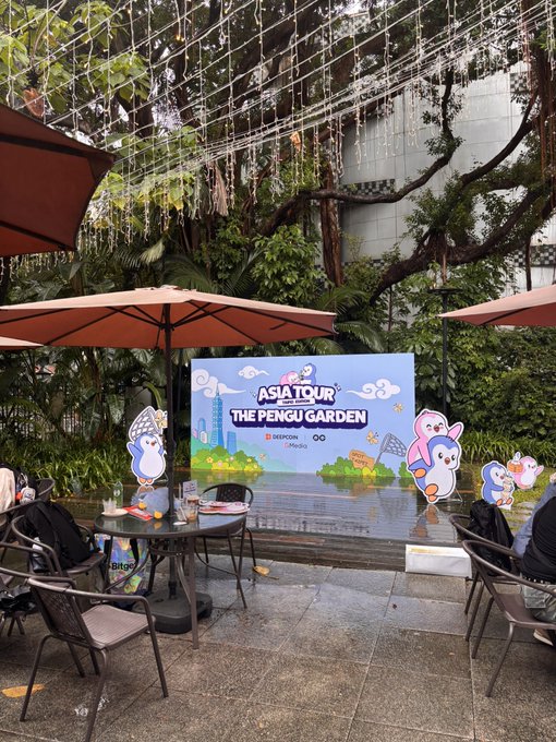 An outdoor venue at the Asia Tour "The Penguin Garden" event, co-hosted by Deepcoin and Media, features a branded stage with penguin mascot cutouts, set up under string lights and orange umbrellas amid lush greenery despite the rainy weather.