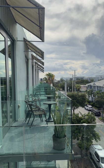 A balcony view from a high-rise building showing outdoor café seating with metal chairs and tables, overlooking a street with palm trees and cars under a cloudy sky.