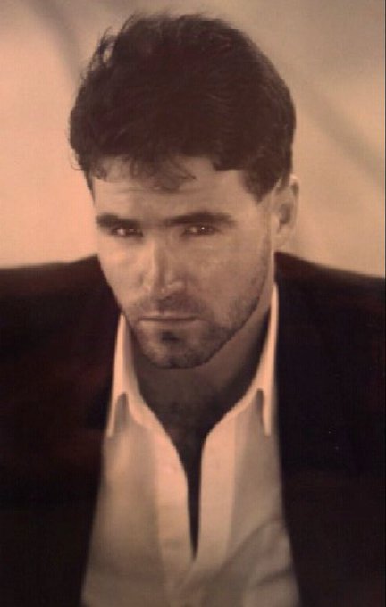 The image features a man wearing a suit jacket, white shirt, and black pants. He has short hair and looks directly at the camera. The photo appears to be from an older time period or vintage style, as indicated by its grainy texture. The caption of the image reads "I have to go back in time 25+ years if I am going to have a LooksMaxxing Showdown with @davidgokhshtein". This suggests that the man is participating in a photo shoot or event where they are trying to recreate an older, vintage look for their images.
