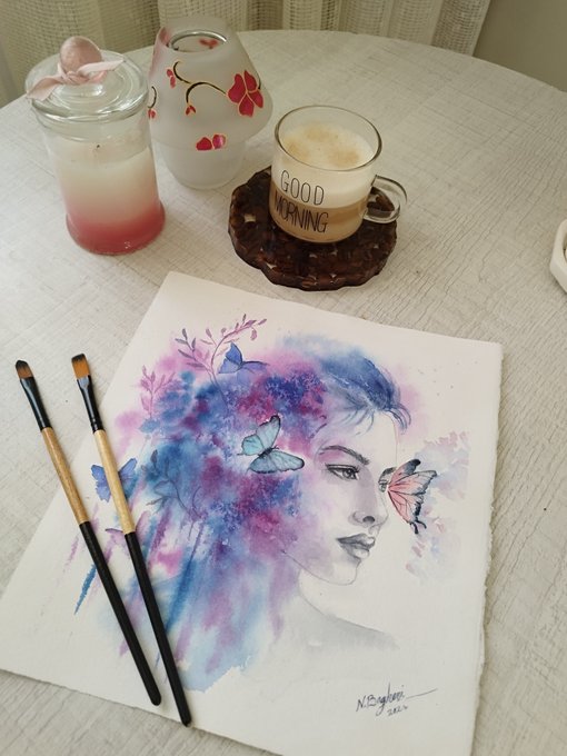 A watercolor painting of a woman surrounded by purple and blue floral elements and butterflies, signed by the artist, displayed on a table alongside paintbrushes and a 'GOOD MORNING' coffee mug.