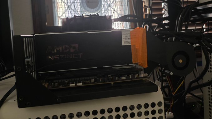 An AMD Instinct GPU sits installed in a computer setup, its bold branding visible on the side of the card. Captured against a window with a fan and cable clutter nearby, the image reflects the excitement of having powerful AI/hpc-grade hardware at one's fingertips — fittingly echoing the post's sentiment about living in an exciting era of accessible high-performance computing.