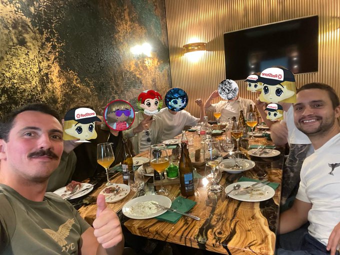 A group photo of men at a dinner table with empty plates and bottles; several faces are covered by stickers including 'RedStore' mascots, Paimon, and a Discord logo.