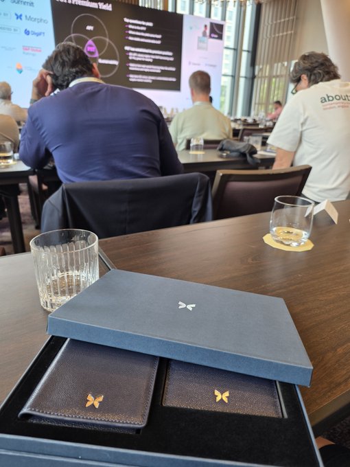 A conference or seminar setting shows attendees seated at tables facing a presentation screen displaying a Venn diagram. In the foreground, a navy blue box with a butterfly logo sits open, revealing two leather card holders also branded with the gold butterfly emblem, alongside a glass of water and a rocks glass with a coaster.