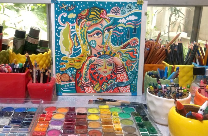A vibrant watercolor painting titled "Daughter of the Sun," featuring stylized faces and flowing figures in bright colors, is displayed surrounded by an artist's tools including colored pencils, markers, and an open watercolor palette, celebrating the recent sale of this piece to collector Robert Rankine.