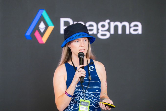 A woman with long blonde hair wearing a blue bucket hat and blue patterned tank top speaks into a microphone at a Pragma event, holding a yellow device.