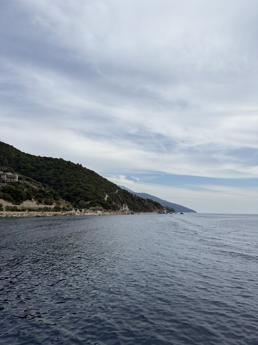 A scenic seascape showing calm blue water with a tree-covered coastline and mountains under a cloudy sky.