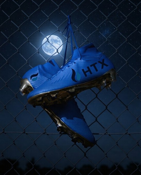 A pair of bright blue HTX-branded cleats hang from a chain-link fence against a starry night sky with a full moon in the background. The nighttime setting and "HODL" reference in the post evoke a sense of holding steady through the night—much like cleats that grip the turf, ready to launch into a legacy run.