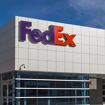 The exterior of a modern building displays the purple and orange FedEx logo on a paneled facade under a blue sky.