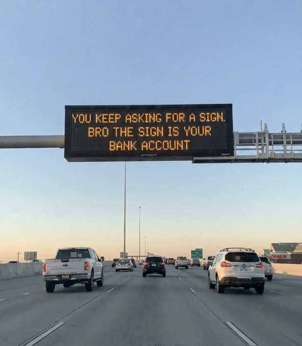 An overhead highway sign reading 'YOU KEEP ASKING FOR A SIGN. BRO THE SIGN IS YOUR BANK ACCOUNT' above traffic on a multi-lane road.