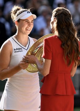 A smiling female tennis player in a white outfit and cap receives a large round championship trophy from a woman in a red dress, capturing the celebratory moment of a Wimbledon victory that matches the post's mention of Noskova's winning payout.
