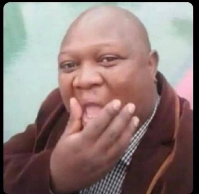 A close-up, slightly blurry photograph shows a bald man with a brown jacket smiling with his hand resting against his face and mouth.