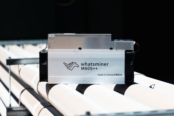A photograph of a Whatsminer M60S++ Bitcoin mining machine mounted on a rack with white pipes in the background.