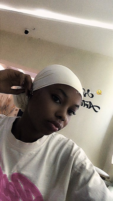 The image features a young woman wearing a white headscarf or hat, possibly a hijab, with her hair styled in an updo. She is posing for a photo while looking directly at the camera. The background of the image shows a wall with graffiti on it and a window that allows natural light to enter the room. There are also some words visible in the image, which appear to be related to the post "GM Web3 friends Happy Wednesday. Certainly goodness and UsDt shall follow us."