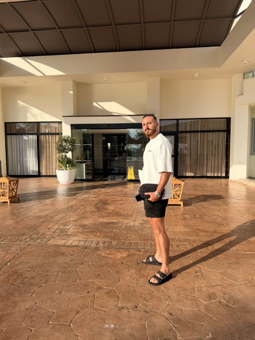 A man in a white t-shirt and black shorts poses for a casual photo on the tiled patio of a modern resort-style building, with a yellow "Caution Wet Floor" sign visible in the background. The relaxed, sun-drenched setting perfectly captures the easygoing "vibe check" mood of the post.