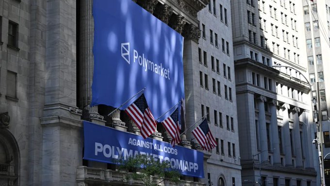 A large blue banner on the facade of a classical stone building displays the Polymarket logo and text reading "AGAINST ALL ODDS POLYMARKET.COM/APP," hung above American flags.