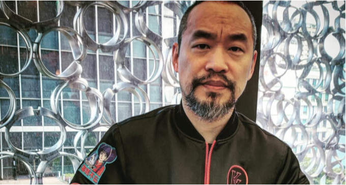 A man with a goatee smiles in front of a decorative window with circular patterns, wearing a black bomber jacket with patches.