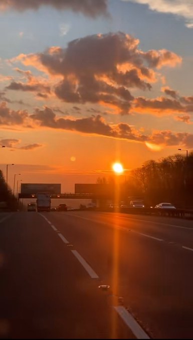 A highway stretches toward a glowing sunset, with dramatic orange clouds and silhouettes of trucks and cars visible in the distance.