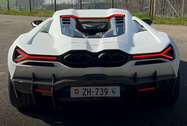 The image features a white Lamborghini sports car with red lights on its rear bumper. The car has a license plate that reads "ZH-739". It appears to be parked, possibly at an event or gathering. The background of the photo is blurred and does not provide any additional information about the location or context.
