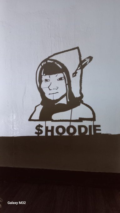 A black and white wall decal/stencil of a hooded figure with a feather holding a pen, with '$HOODIE' written below it, photographed on a Samsung Galaxy M32.
