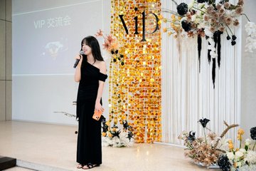 The image features a woman standing on stage wearing a black dress with gold accents. She is holding an orange object, possibly a microphone or a microphone stand, as she speaks into it. The background of the scene includes a large screen displaying text that reads "VIP" and "bitget". There are also flowers visible in the image, adding to the overall ambiance of the event.