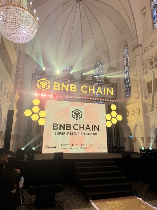 An interior view of a grand hall with a large illuminated stage setup displaying the BNB Chain logo for the 'Super Meetup Singapore', featuring bright stage lights and a large chandelier overhead.