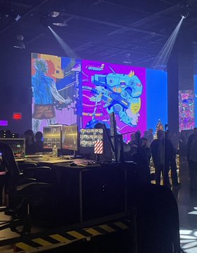 The image shows a large screen displaying Beeple's "Ponzinomics" artwork at what appears to be his studio, featuring his signature colorful, surreal digital art style with intricate mechanical and figurative designs. The scene captures the studio environment with workstations, monitors, and people gathered, complementing the post about visiting Beeple Studios in 2023.