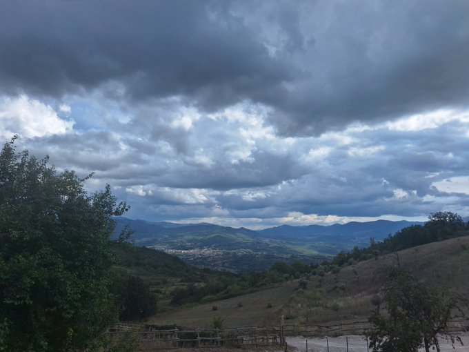 A scenic landscape view showing rolling green hills, distant mountains, and a dramatic sky filled with dark, heavy clouds with patches of blue peeking through.