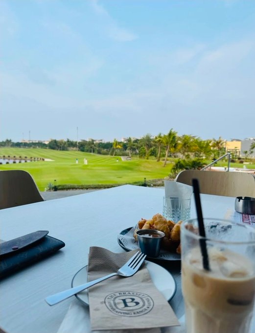 A restaurant table setting with a view of a green golf course, featuring a plate of fried food, a fork on a 'B' branded napkin, and an iced coffee beverage under a blue sky.